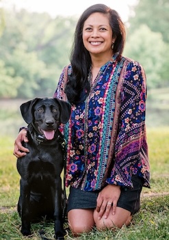 Anne Umali smiles while wearing a multicolored tunic and kneels next to a young Black Labrador. 