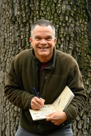 John Muir Laws smiles while nature journaling in front of a large tree