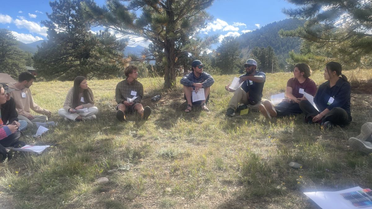 A group sits in a circle in a shaded outdoor gathering. Photo credit: Katie Navin