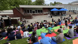 Large group of students seated on a down-sloping lawn facing a paved stage in front of school buildings