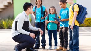 Teacher crouching next to four elementary aged students who are smiling and wearing backpacks.