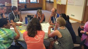 Group of adults with thoughtful expressions gathered around a round table with markers and chart paper. 