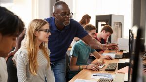 Teenage students working at row of computers. Teacher appears to be helping one student, as they look at the monitor together. 