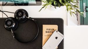 Photo of a desk with a keyboard, mouse, phone, notebook, headphones, computer screen, plant, and book on top