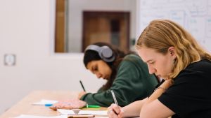 Two students work intently. One listens to something through headphones. 