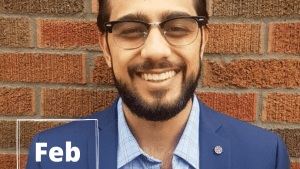 Brown-haired person smiling at the camera while in front of a brick background. On the lower left corner is a white rectangle with text inside that says, "Feb 21."