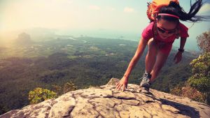 Hiker climbing rock on mountain peak. Lush green landscape below.