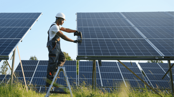 A person in a white hard hat stands between two solar panels, working on the one to the right