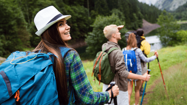 A group of young hikers wearing backpacks and holding trekking poles walk along a grassy trail near a forest and lake