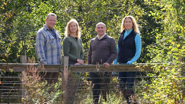 Four light-skinned people stand on a boardwalk with a wooden rail. Surrounding them is green foliage from herbaceous plants, shrubs, and trees.