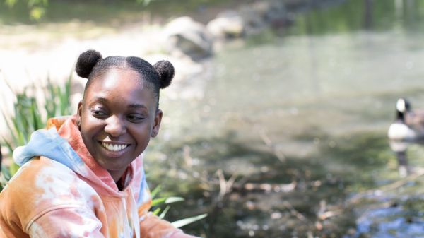 A smiling Black woman looks off to the side while near a duck pond