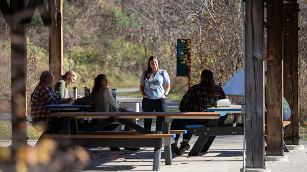 A group of people at picnic tables underneath a large outdoor shelter. In the middle, one person in a light-blue shirt stands and faces the others. In the background, mostly leafless trees with a few green conifers.