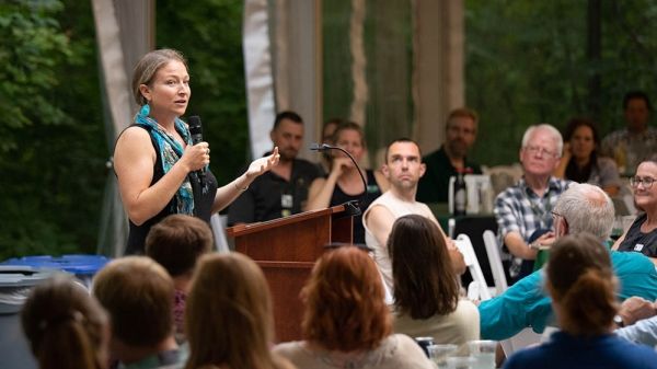 A person stands at a podium and holds a microphone, addressing a crowd. In the foreground and background, the seated crowd listens.