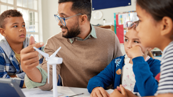 A teacher sits at a table surrounded by young students, pointing to a miniature wind turbine