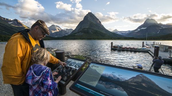 An adult an a child read an interpretive sign next to a mountain range and a body of water An adult an a child read an interpretive sign next to a mountain range and a body of water