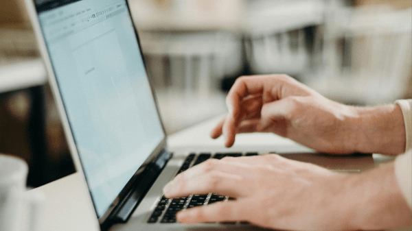 Two hands type on a laptop on a white table; white chairs can be seen in the blurred background