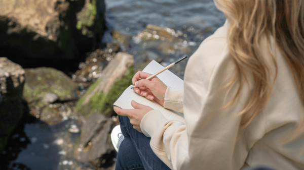 Nature Journaling Rocks with algae sit in water to the left; to the right a person is sitting, holding a journal in their lap with a pencil in their right hand