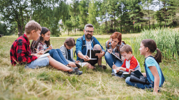 Two adults sit in grass, one holding a miniature wind turbine, surrounded by children and all in a grassy field with trees further in the background