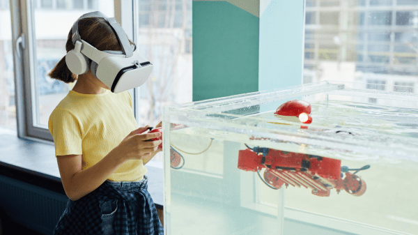 A child wearing a VR headset stands to the left of a water tank with a controller in their hands; the water tank contains a small red robot