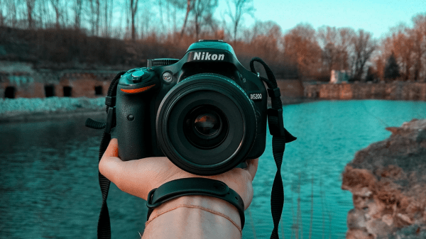 A hand holds out a camera facing the viewer, beyond it is a river