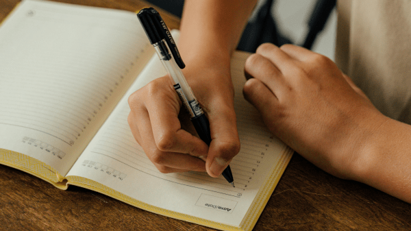 Top Action Tips Two hands are over a notebook on a brown desk; one hand holds a pen ready to write, the other the bottom corner of the notebook