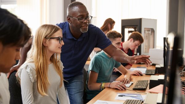 Teenage students working at row of computers. Teacher appears to be helping one student, as they look at the monitor together.