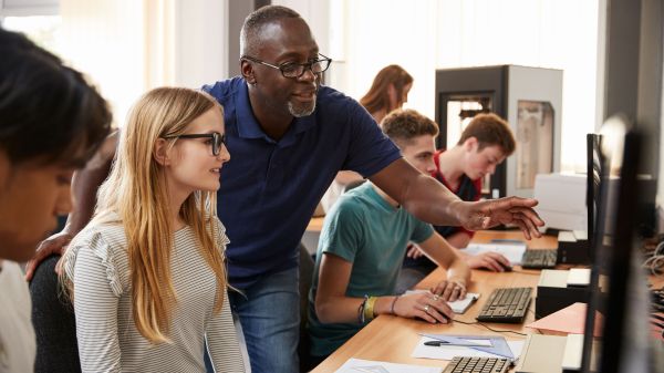 Teenage students working at row of computers. Teacher appears to be helping one student, as they look at the monitor together. 