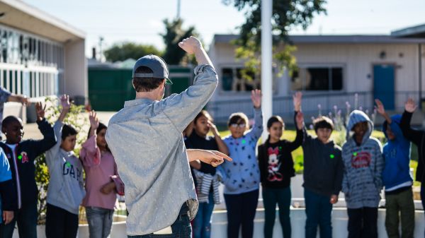 Teacher standing, hand raised, infront of a group of students in a outdoor learning space.