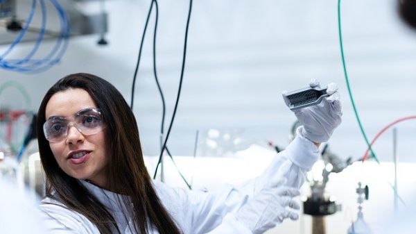 A woman wearing safety glasses in a lab setting with wires in the background, holding a metallic bracket