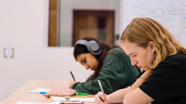 Two students work intently. One listens to something through headphones. 