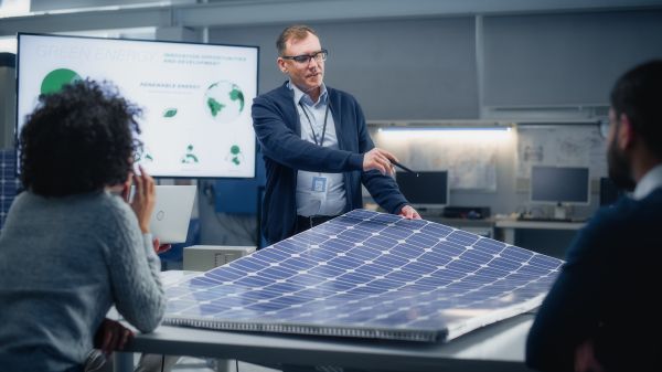 Three people are arranged around a solar panel; one person is standing and holding part of the panel with a presentation board behind them
