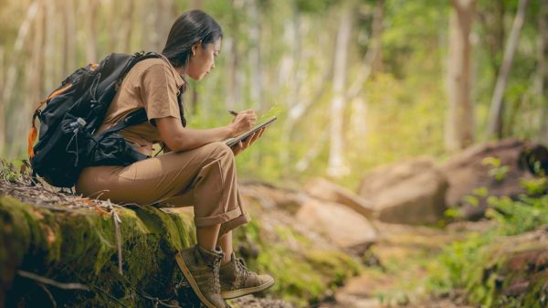 Person wearing backpack, sitting on mossy stones in forest to take some notes.