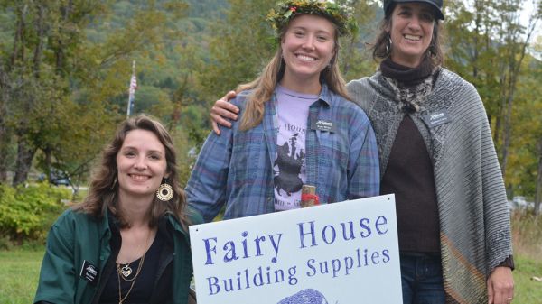 Staff of The Nature Museum in Grafton, Vt. Three light-skinned and long-haired people smile behind a yard sign that reads "Fair House Building Supplies." The person to the left crouches, and the two people to the right stand. Behind them, green grass and trees in late summer foliage.