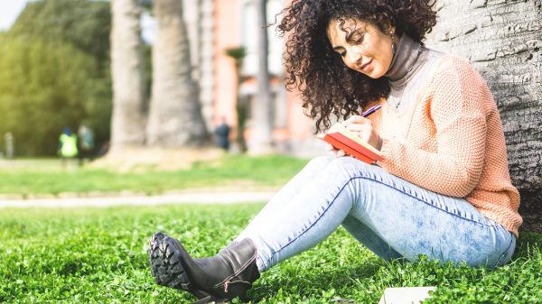 Person sitting on grass against a tree, writing in small journal.