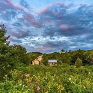 A building stands in the distance, surrounded by a prairie in the foreground, forested hills behind, and a stunning blue sky above.