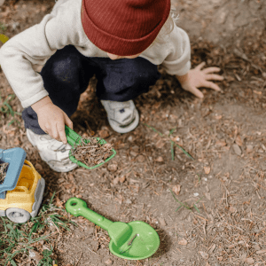 A child digs in the dirt with a green plastic shovel, below them is a plastic truck and another shovel