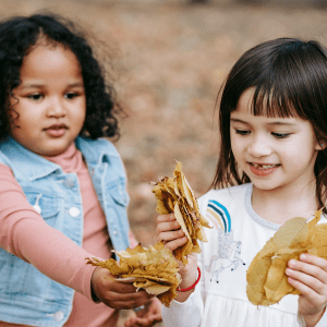 Two children hold clumps of brownish orange leaves, behind them is a blurred background where leaves can be seen on the ground