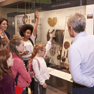 A group of five children stand in front of an adult, three raising their hands; to the right is an adult facing them; behind all of them is a display of fossils