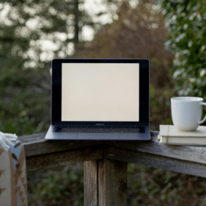 An open laptop sits on a patio fence between a blanket and a coffee mug; behind it is a forest of trees