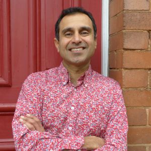 A smiling Vik Mohan wearing a red long-sleeve button-up shirt and standing in front of a red door