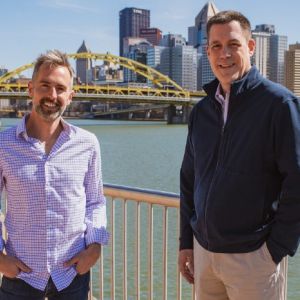 Ryan Rydzewski and Gregg Behr stand in front of a yellow bridge in Pittsburgh, Pennsylvaina.