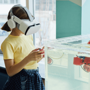 A child wearing a VR headset stands to the left of a water tank with a controller in their hands; the water tank contains a small red robot