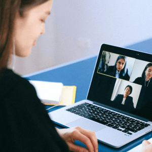 A person sits to the left with an open laptop on a blue desk in front of them; the laptop is open to a video call with three people