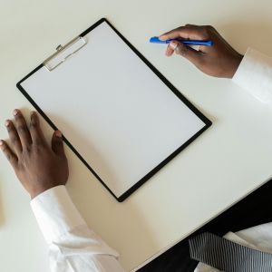A clipboard on a white desk with two hands next to it, one holding a pen