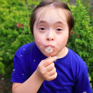 A child outside blowing dandelion seeds in front of greenery