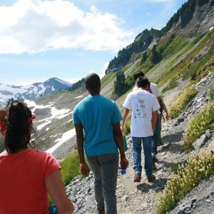 A line of four teens hiking on a mountain trail. Snow-capped mountains in the distance.