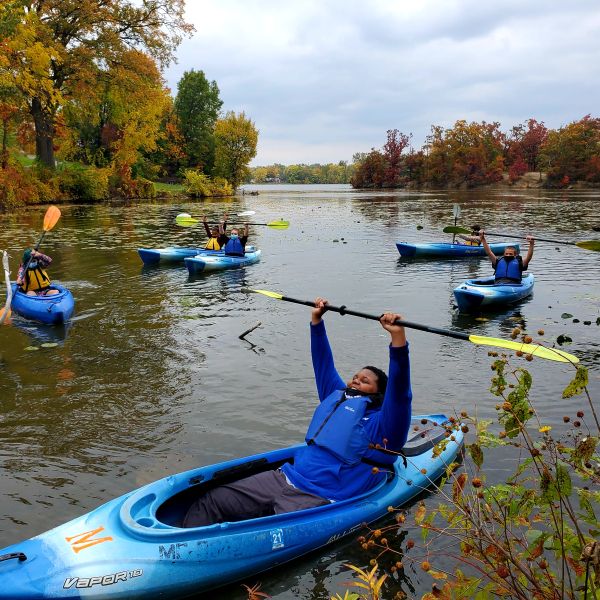 Seven youth in kayaks on lake pose raising paddles above heads and smiling