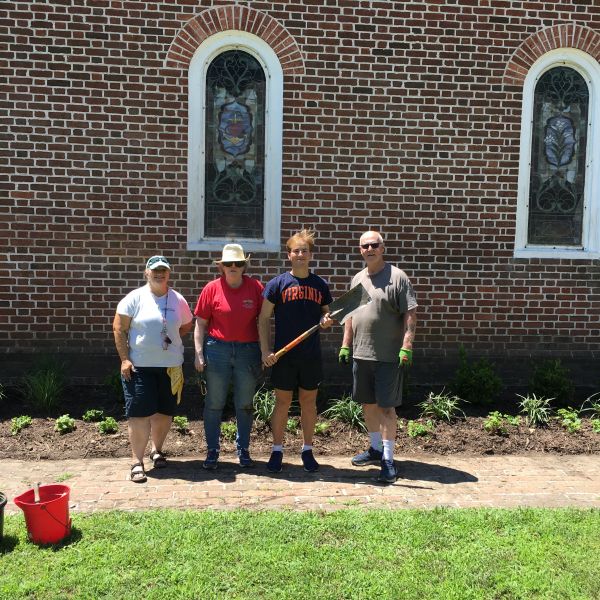 A group of four people stand, facing the camera, in front of a brick building with a garden bed.